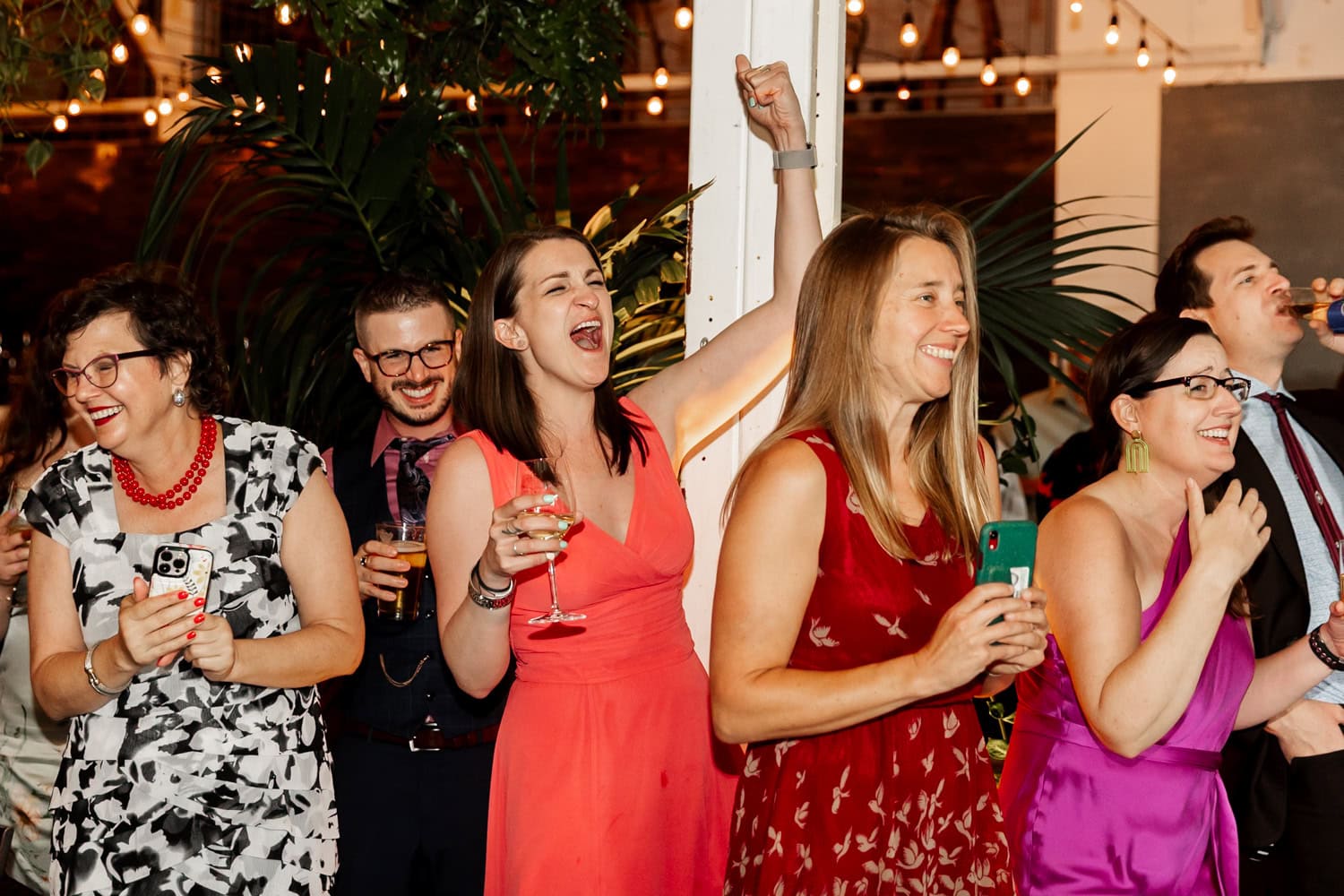 A lively wedding reception at Coopers Hall with guests in colorful outfits, cheering and celebrating indoors while holding drinks under festive lighting.