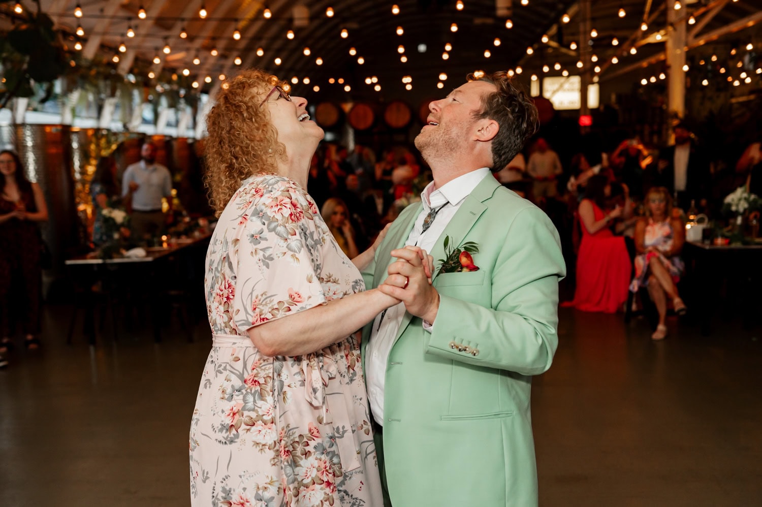 A couple in floral and mint outfits dances joyfully under string lights at a wedding reception at Coopers Hall.