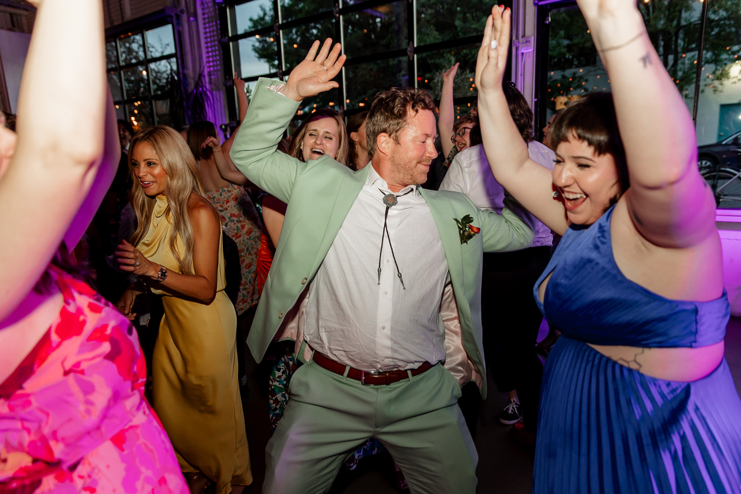 A group of people dancing energetically at a wedding reception at Coopers Hall, surrounded by colorful lights and showing joyful expressions.