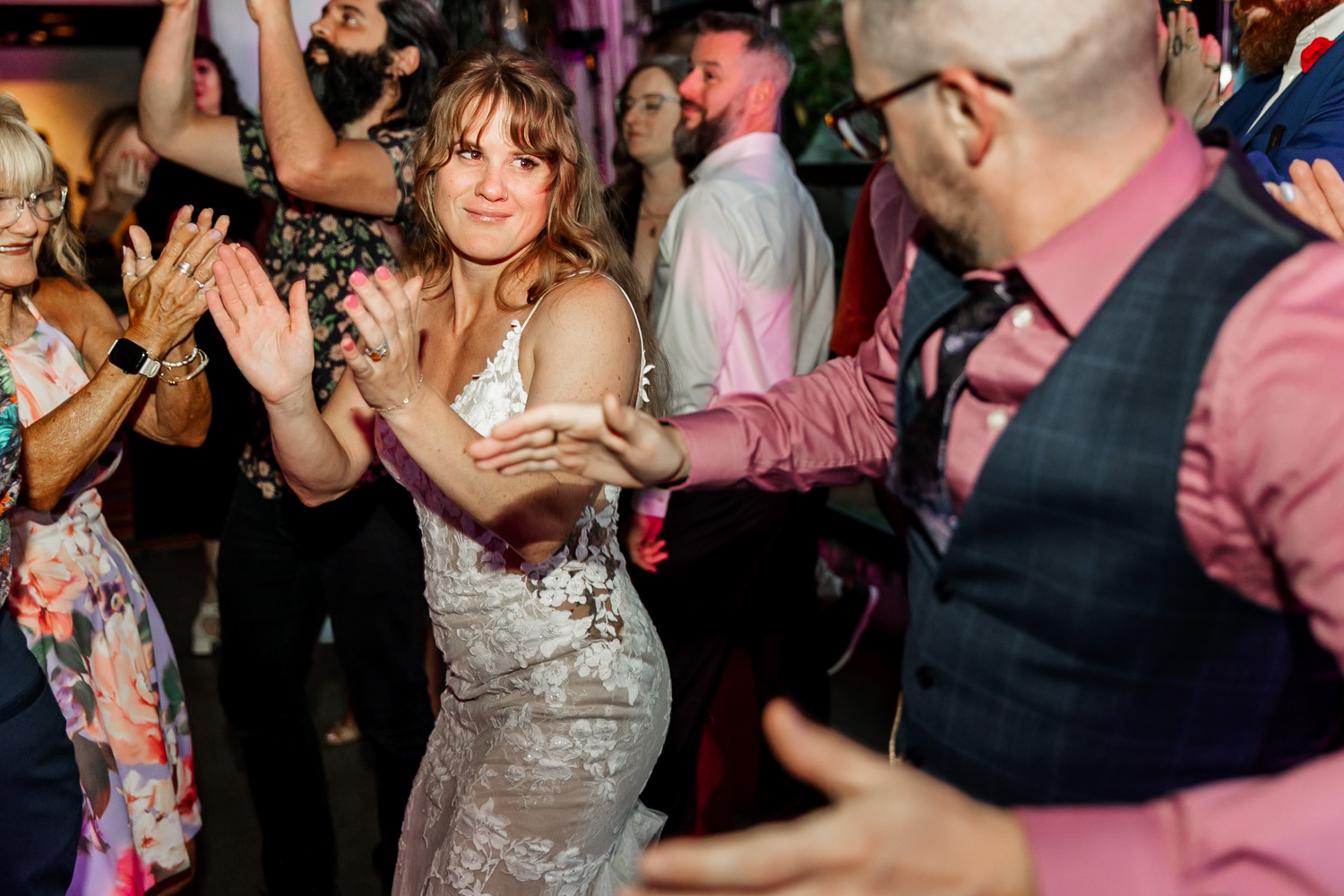 A bride in a lace dress dances joyfully at Coopers Hall during a wedding reception, surrounded by people clapping and joining in the celebration.