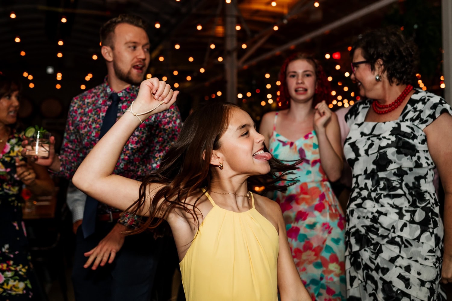 A girl in a yellow dress dances joyfully at a wedding reception, surrounded by guests in colorful outfits. Overhead, string lights cast a warm glow, adding to the festive atmosphere at Coopers Hall.