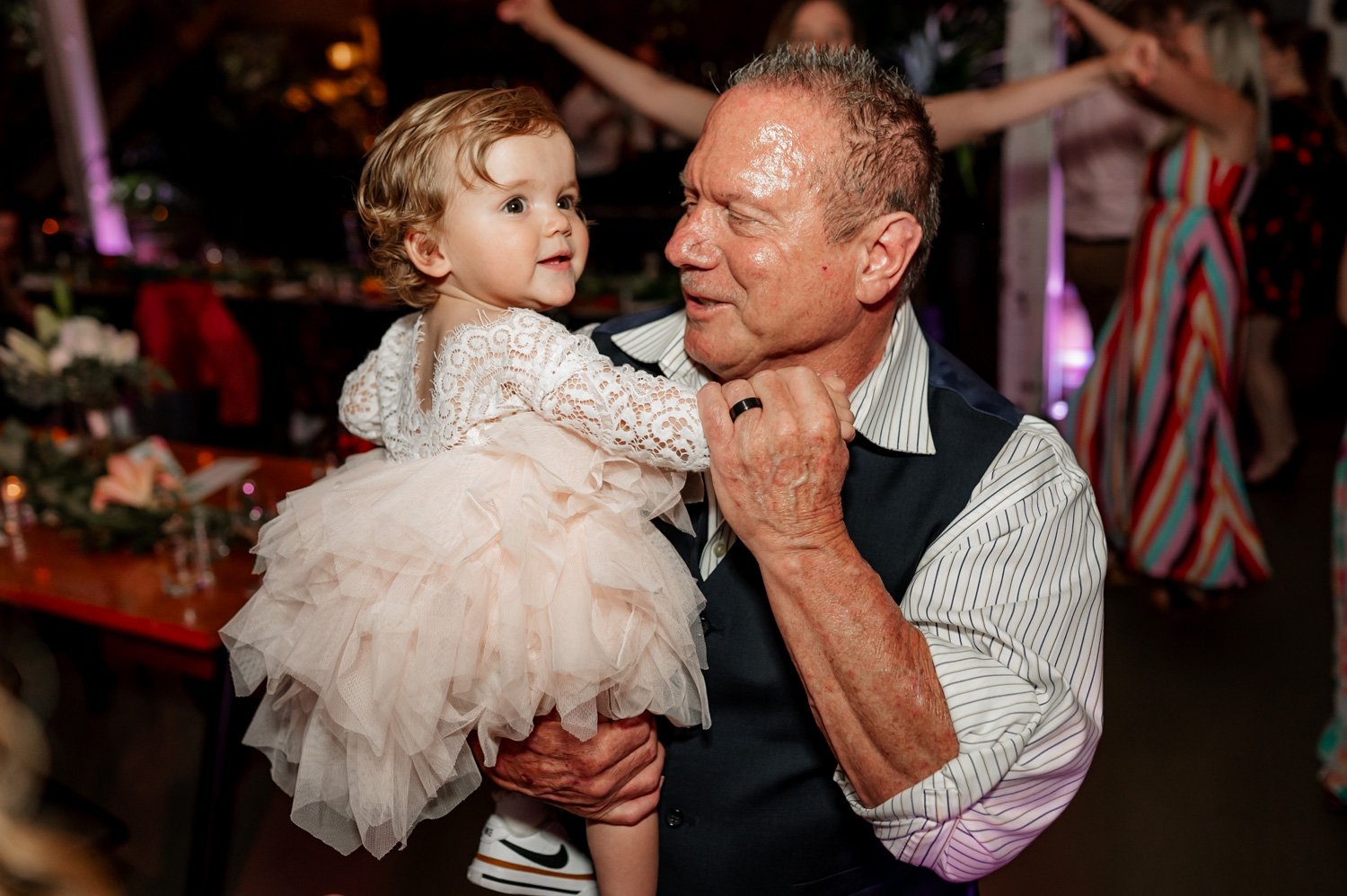 An elderly man holds a toddler in a white dress at Coopers Hall, with guests dancing joyfully in the background at a wedding reception.