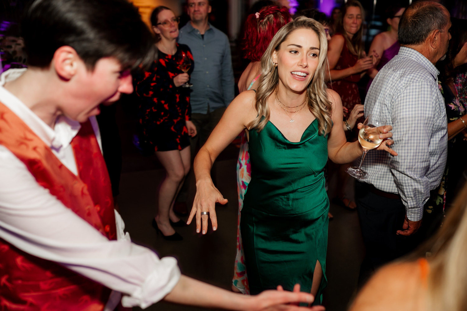 A lively wedding reception with guests dancing energetically; a woman in a green dress is prominently holding a wine glass in the foreground at Coopers Hall.