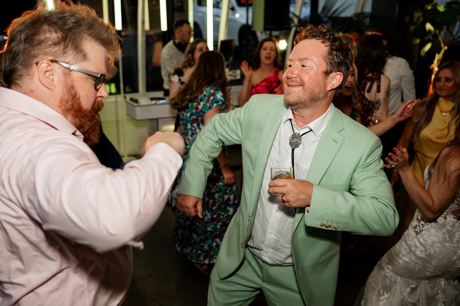 Two men energetically dancing at a wedding reception, with guests in vibrant clothing surrounding them at Coopers Hall.