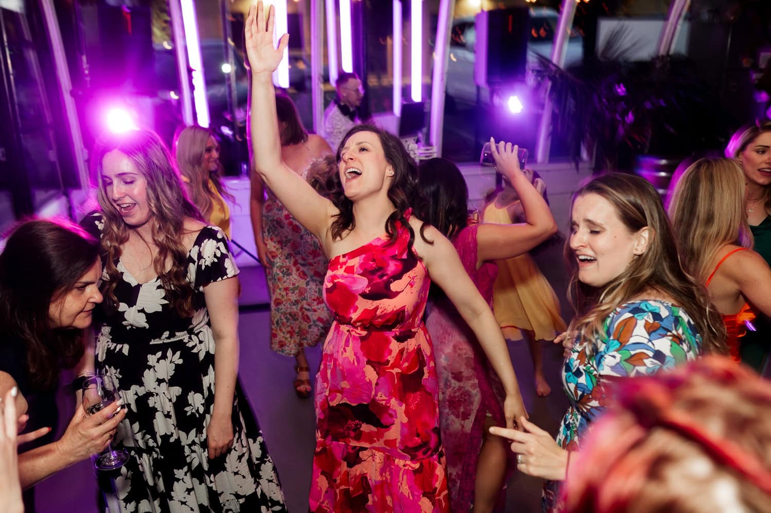 A group of women dance joyfully at a wedding reception with colorful lights swirling in the background.