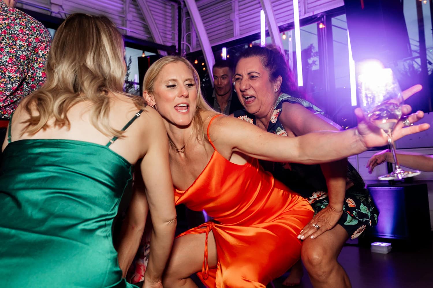 Women in colorful dresses dance energetically at a wedding reception at Coopers Hall, holding drinks and smiling.