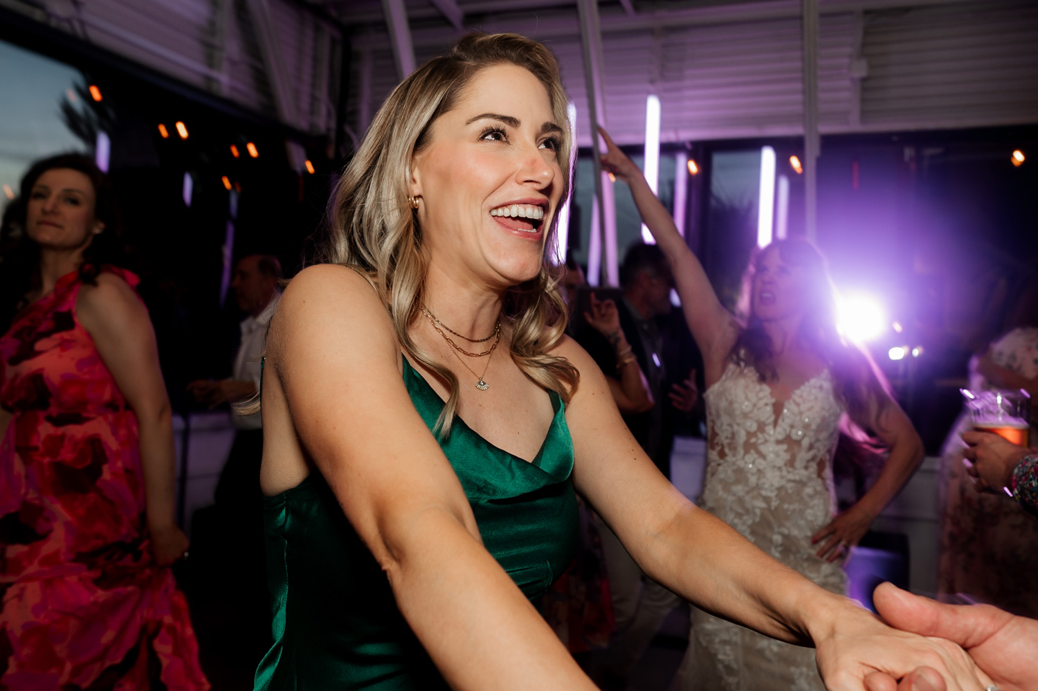 A woman in a green dress laughs and dances at a wedding reception, surrounded by guests in colorful outfits.