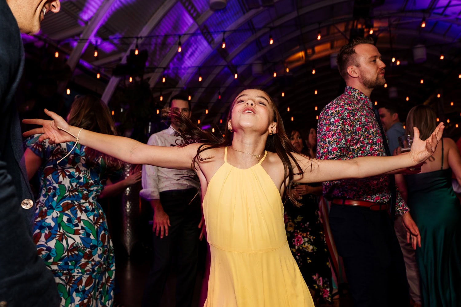 A young girl in a yellow dress dances joyfully at a wedding reception at Coopers Hall, with people and colorful lights in the background.