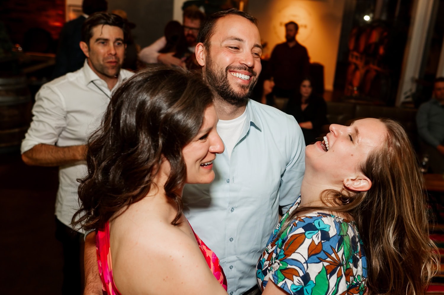 Three people laughing and embracing at a wedding reception at Coopers Hall, with guests mingling and enjoying the lively atmosphere in the background.