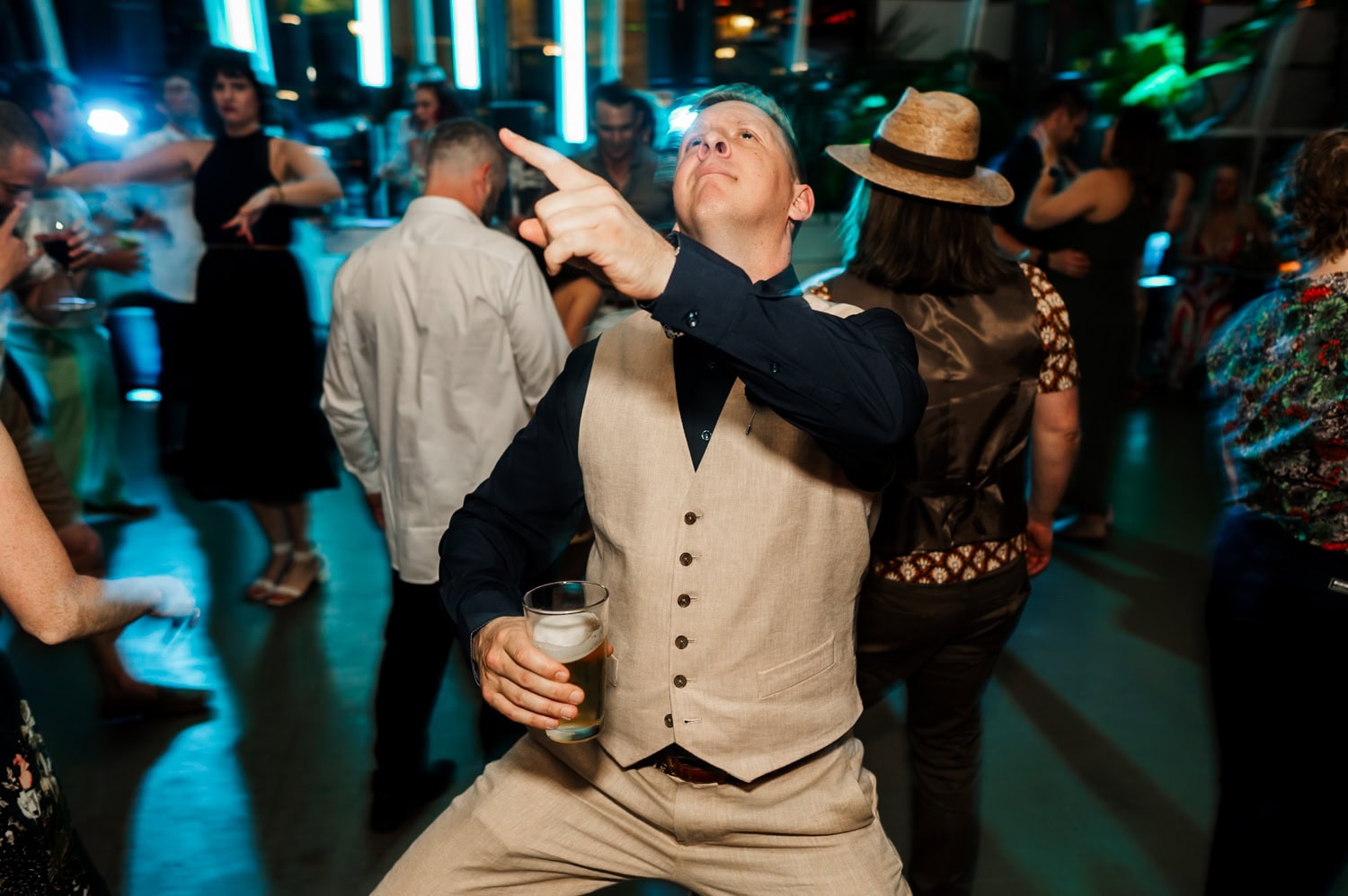 A man in a beige vest energetically dances with a drink in his hand at a wedding reception at Coopers Hall, surrounded by attendees smiling and enjoying the lively atmosphere.