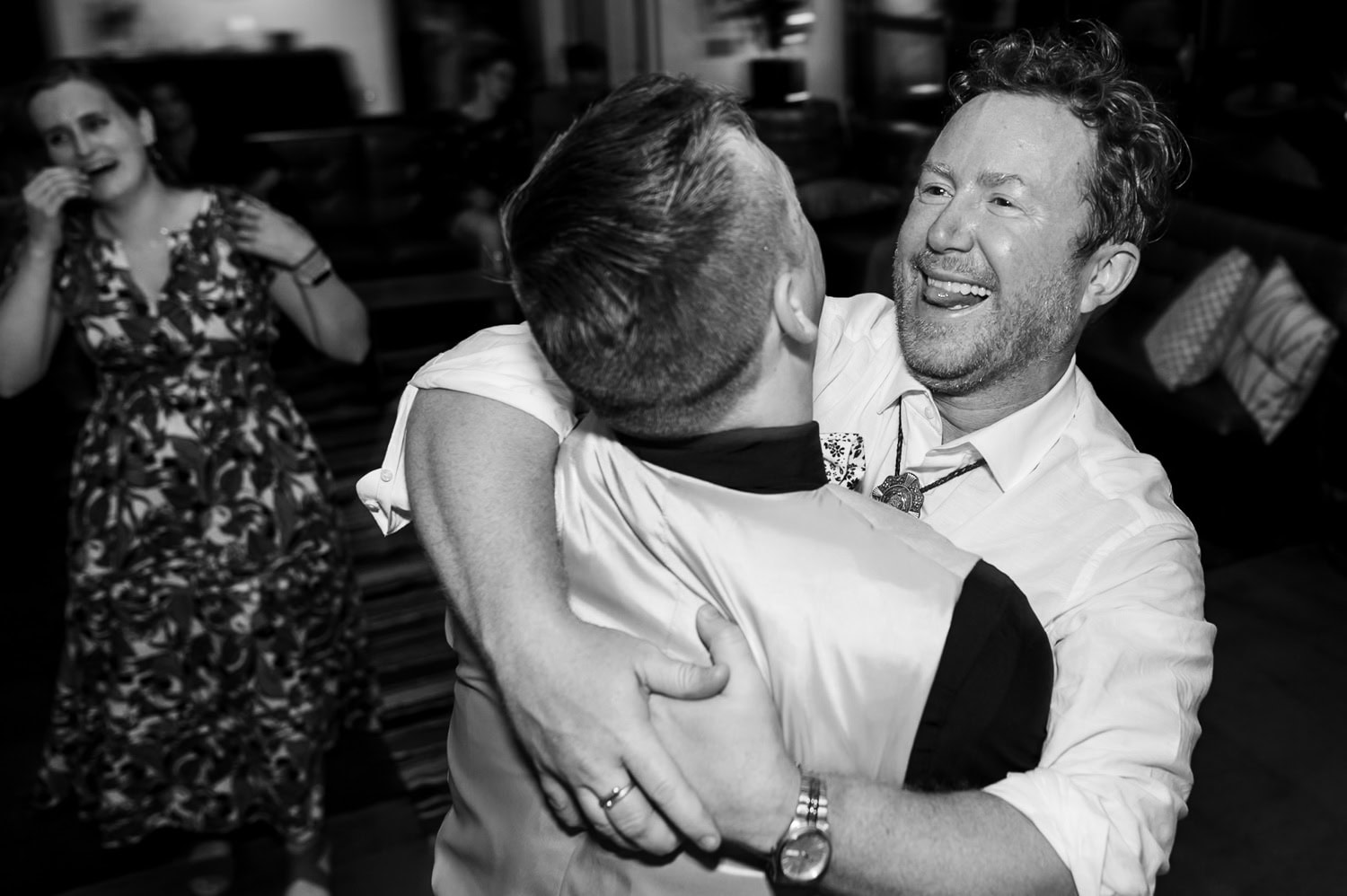 Two people dancing closely and smiling, with a third person watching in the background at Coopers Hall during an indoor wedding reception. The black and white photo captures the joyful atmosphere.