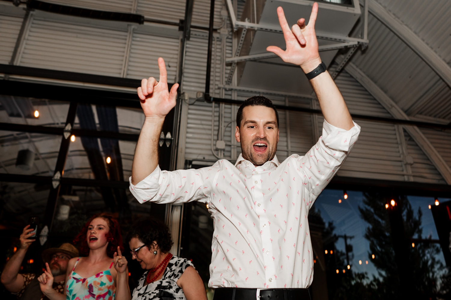 Man in a white shirt joyfully raises his hands at a wedding reception at Coopers Hall, with people and string lights visible in the background.