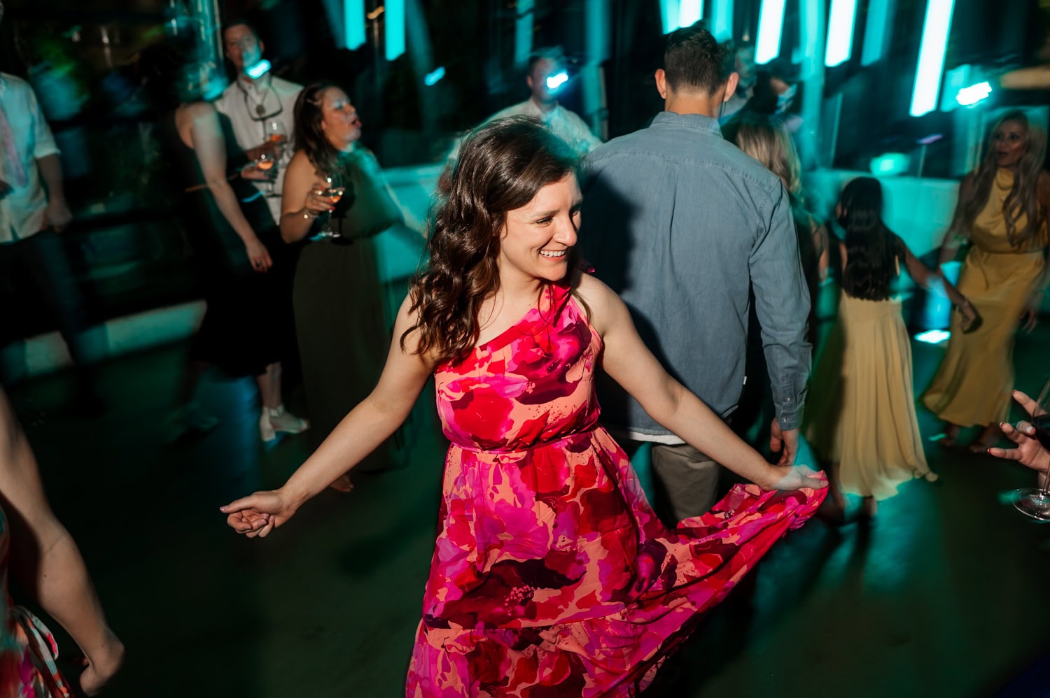 A woman in a vibrant floral dress joyfully dances at a wedding reception at Coopers Hall, surrounded by lively guests in the background.