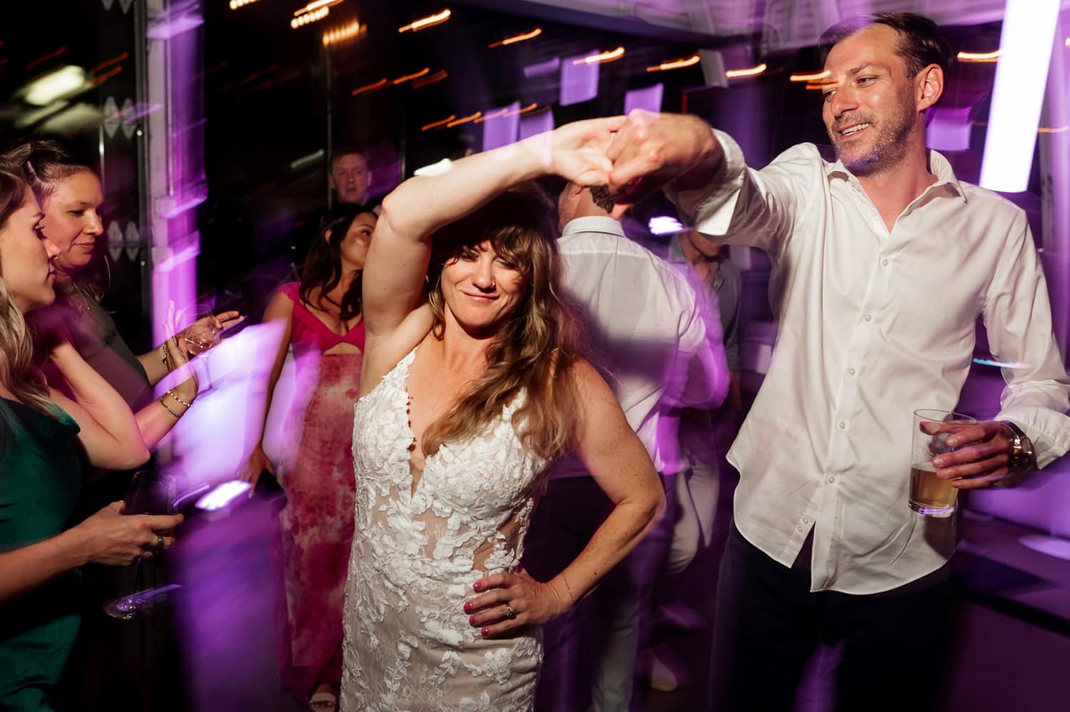 A couple dances joyfully under purple lights at Coopers Hall, surrounded by smiling guests at a lively wedding reception.