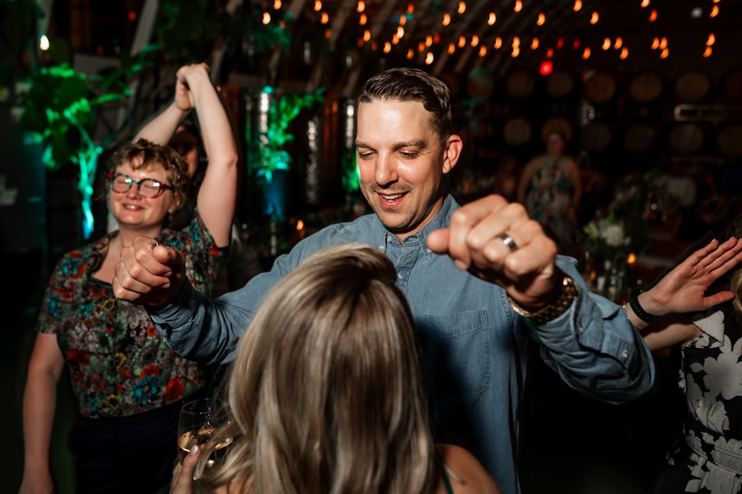 A group of people dancing energetically at a wedding reception in a warmly lit venue adorned with string lights and green decorations, capturing the lively atmosphere at Coopers Hall.