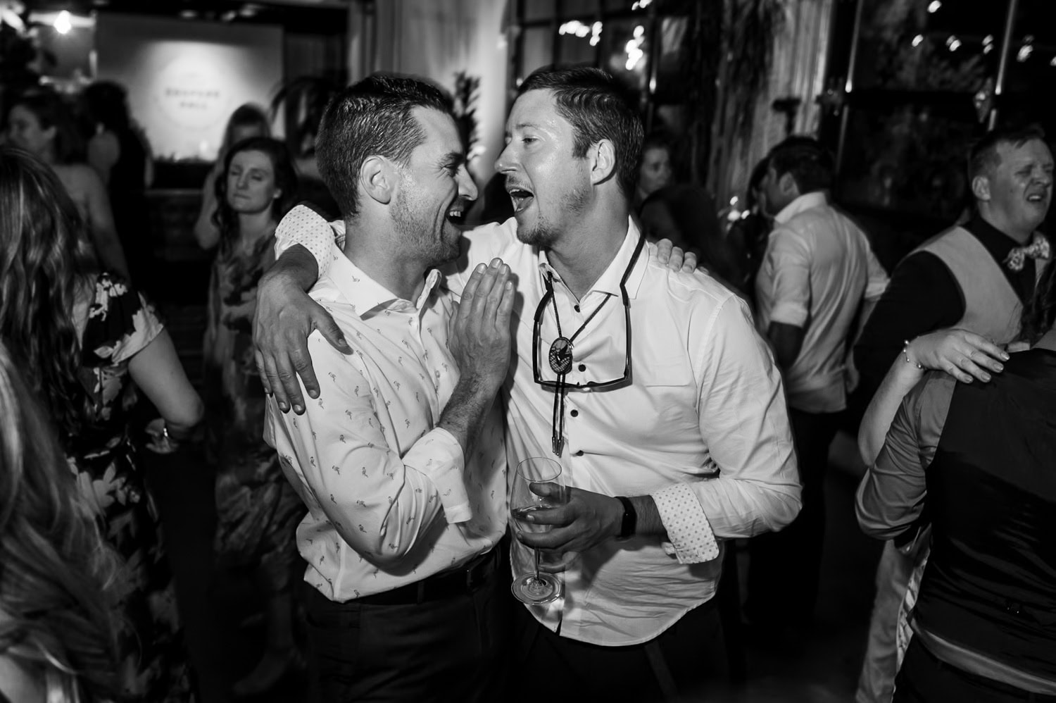 Two men laughing and embracing at a lively wedding reception, with a crowded dance floor visible in the background at Coopers Hall.