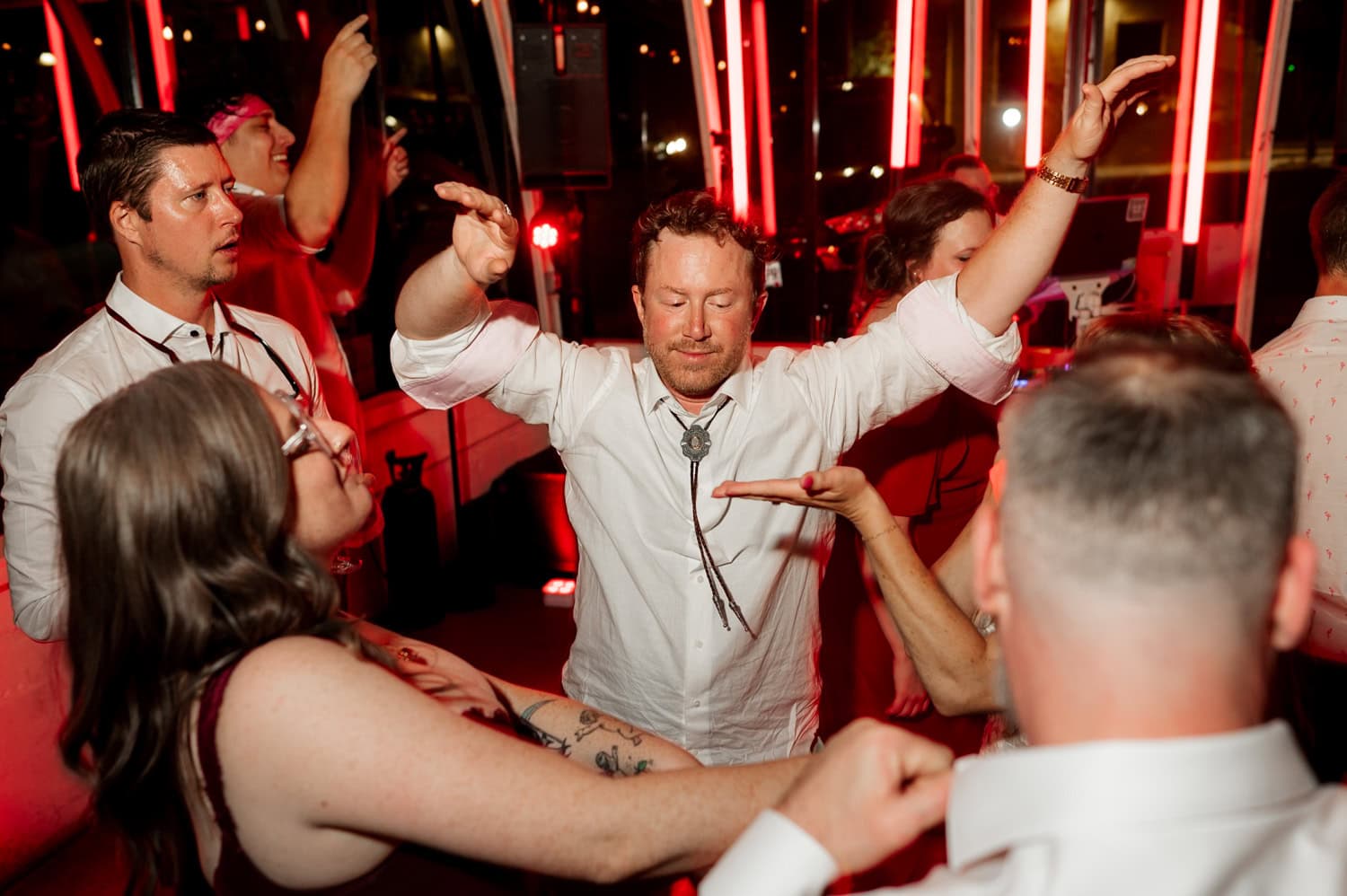 A lively scene of people energetically dancing at a wedding reception in Coopers Hall, illuminated by red lights and dressed in white attire.