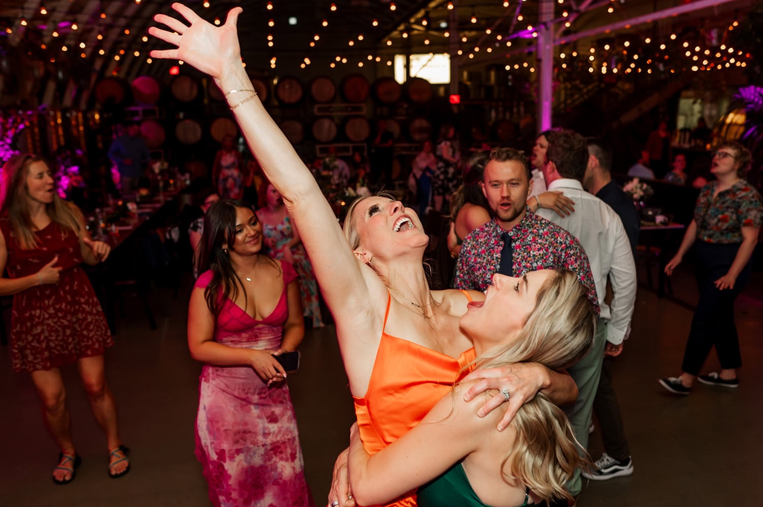 Two people dancing joyfully at a wedding reception, with one raising their arm. Other guests dance and mingle in the warmly lit venue decorated with string lights.