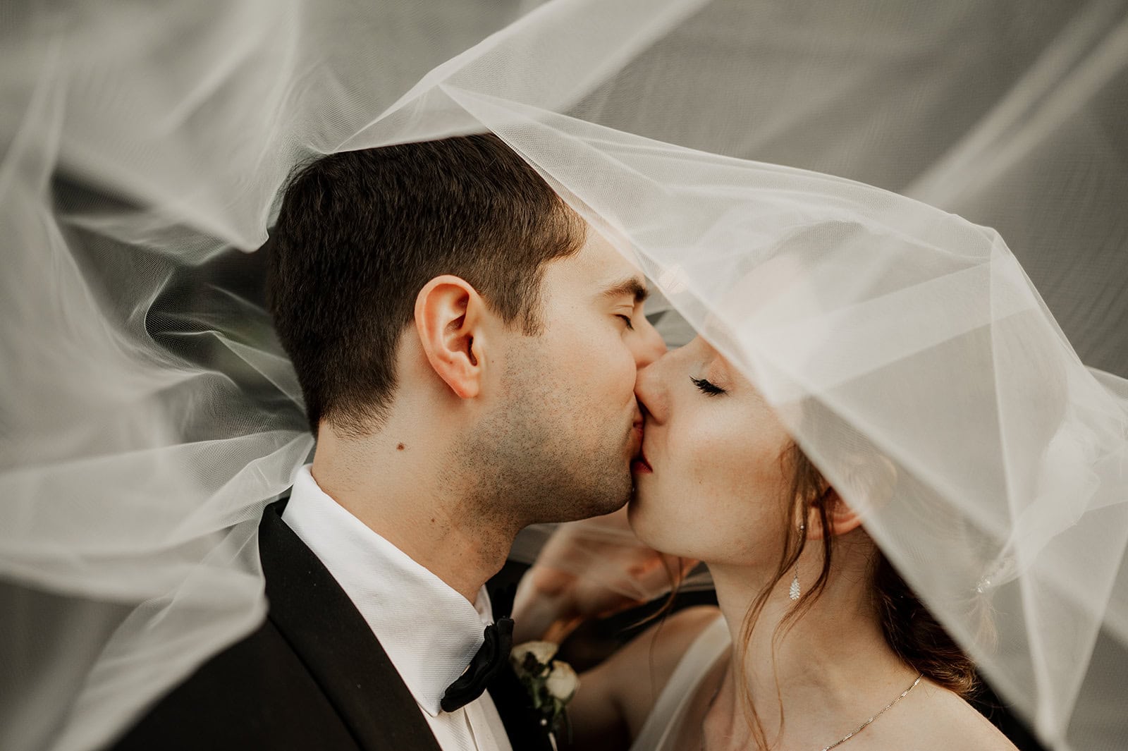 A bride and groom kiss beneath a sheer white veil on their wedding day at The Evergreen in Portland, Oregon, with soft natural light illuminating their faces.