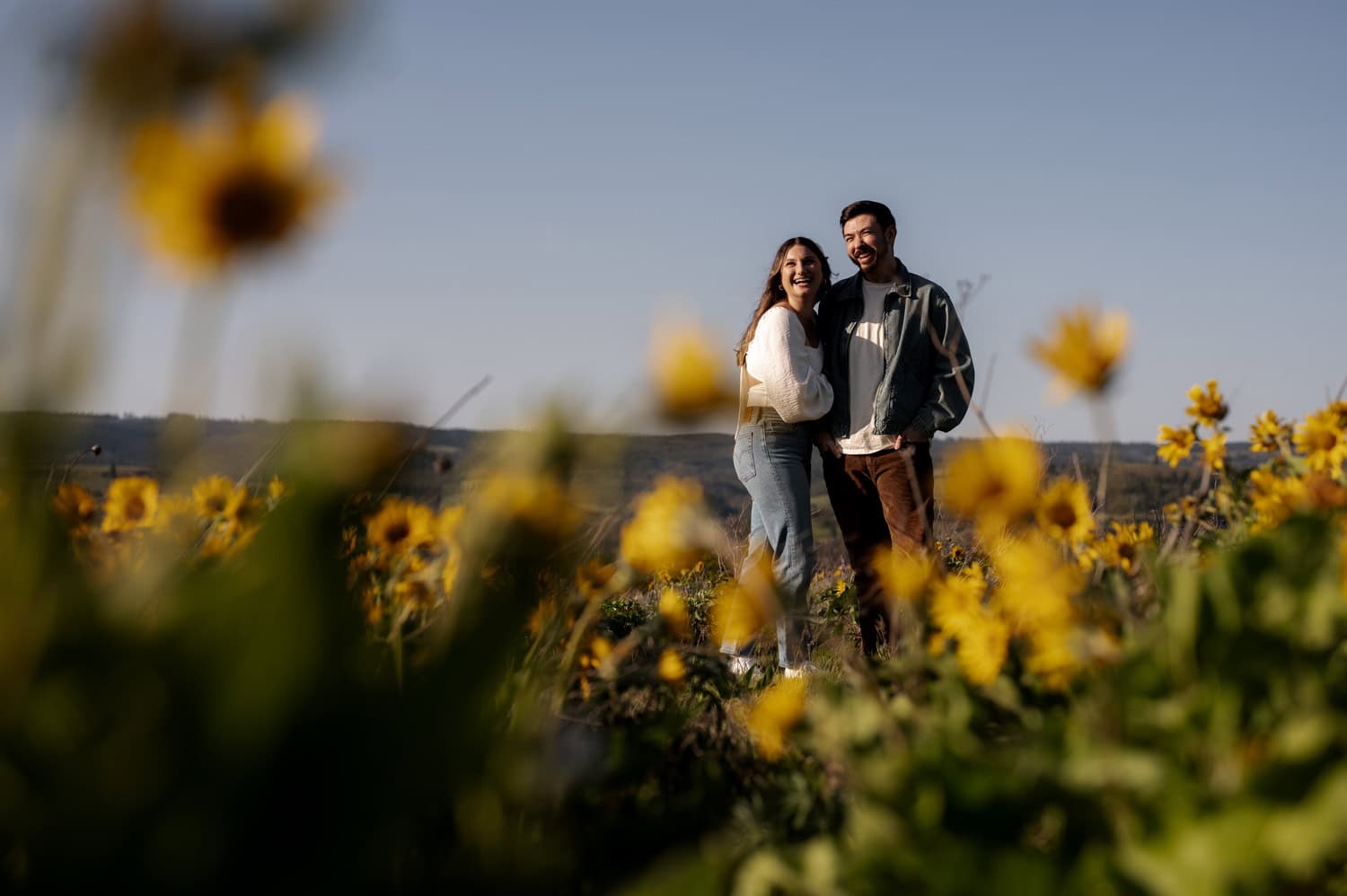A smiling couple stands together in a field of colorful wildflowers under a clear blue sky at Rowena Crest, with green hills in the background.