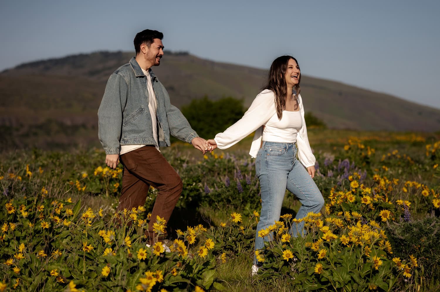 A smiling couple holds hands while walking through a field of yellow wildflowers at Rowena Crest, with rolling hills in the background during their engagement session.