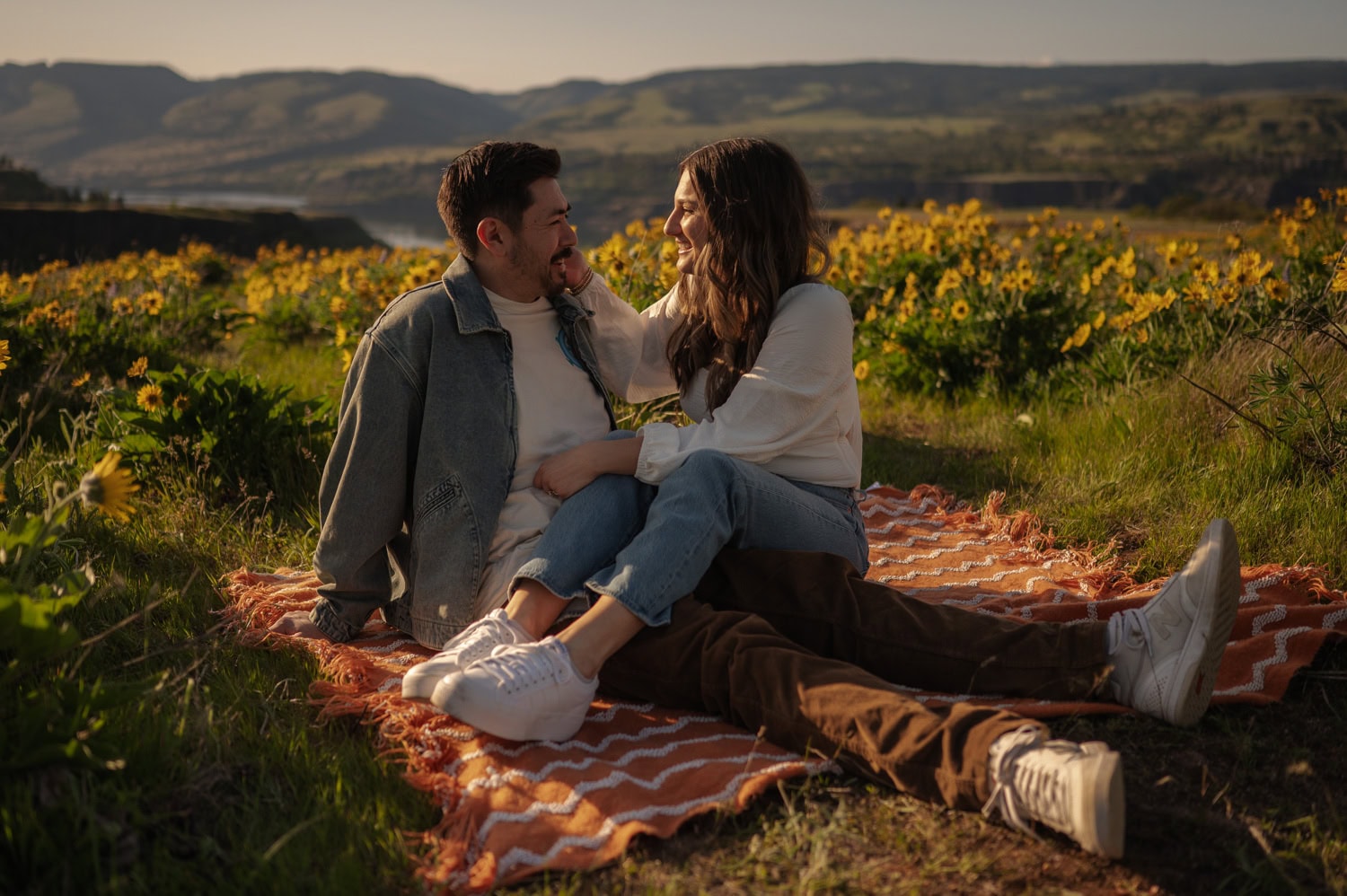 A couple sits close together on a blanket in a sunny field of wildflowers at Rowena Crest, smiling at each other during their engagement session.