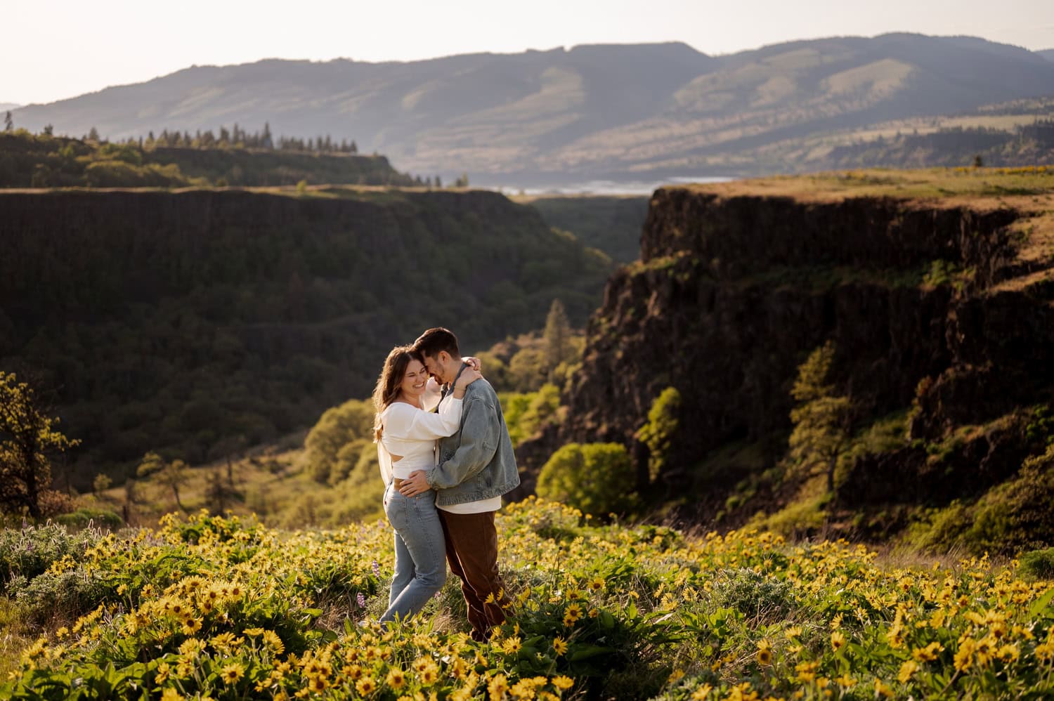 A couple embraces in a field of wildflowers at Rowena Crest, with mountains visible in the background during their engagement session.