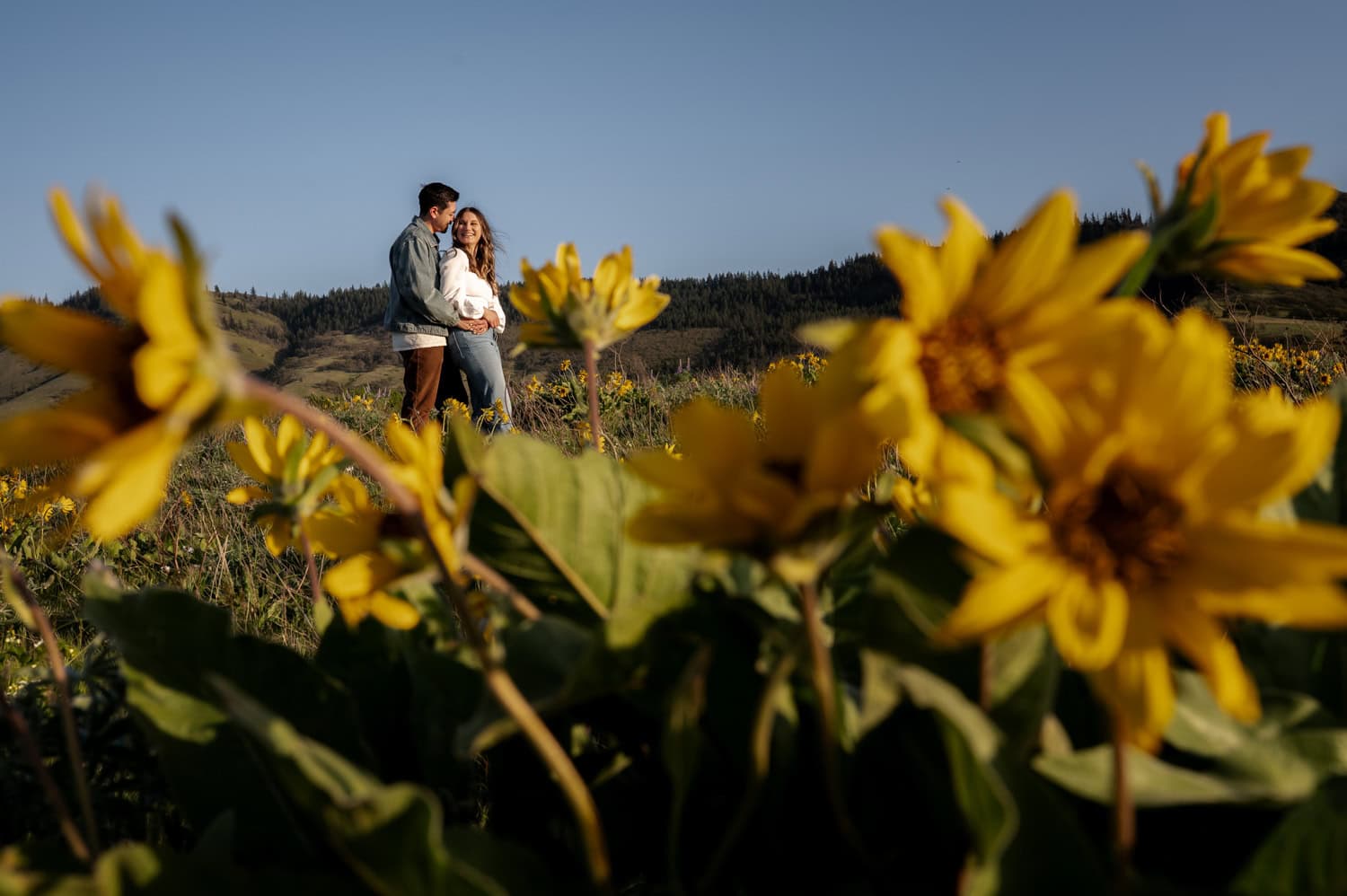 A couple stands together in a field of wildflowers at Rowena Crest, surrounded by rolling hills and trees under a clear sky.