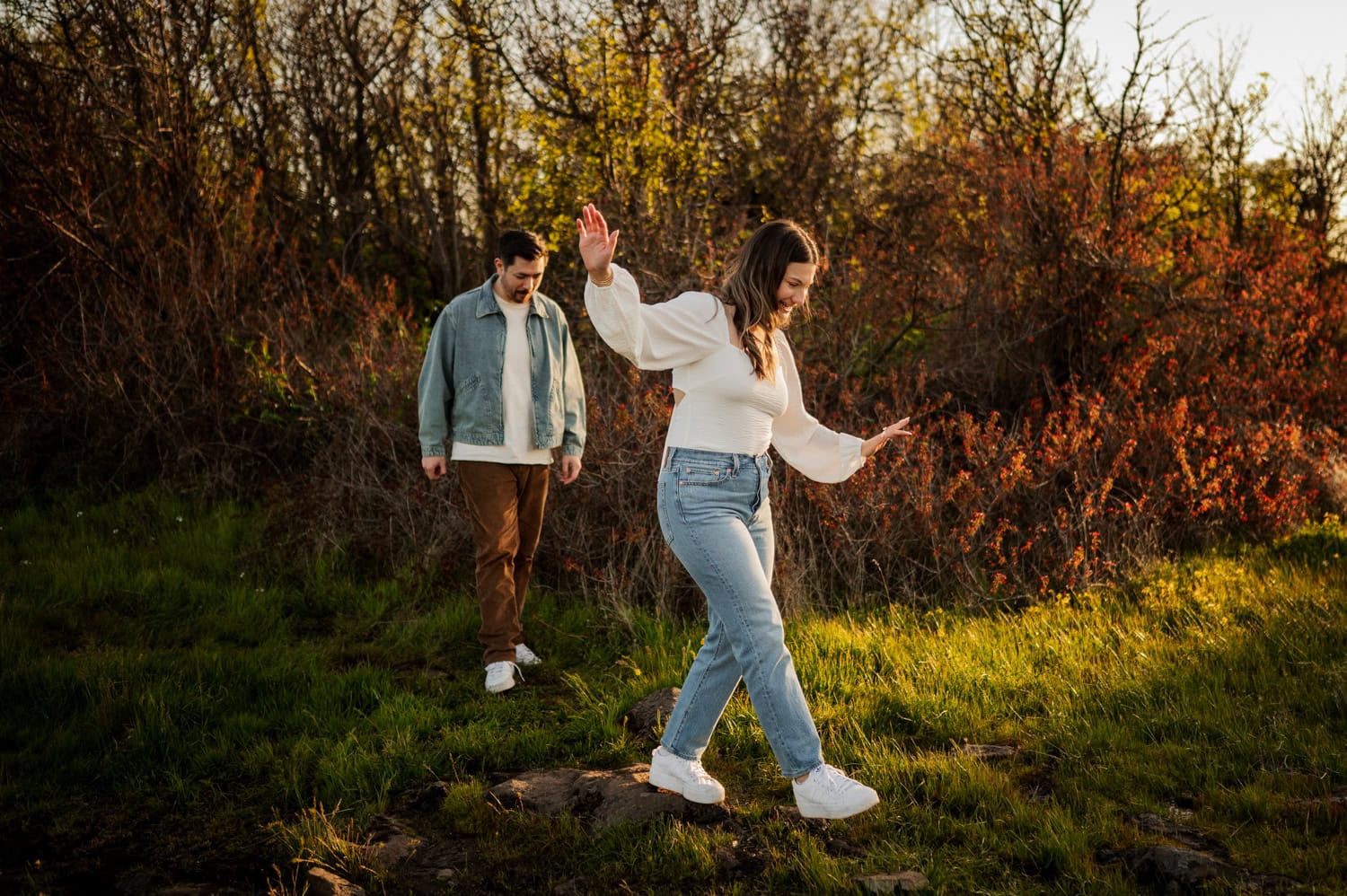 A woman balances on rocks as a man follows behind her in a grassy, sunlit field filled with blooming wildflowers at Rowena Crest during their engagement session.