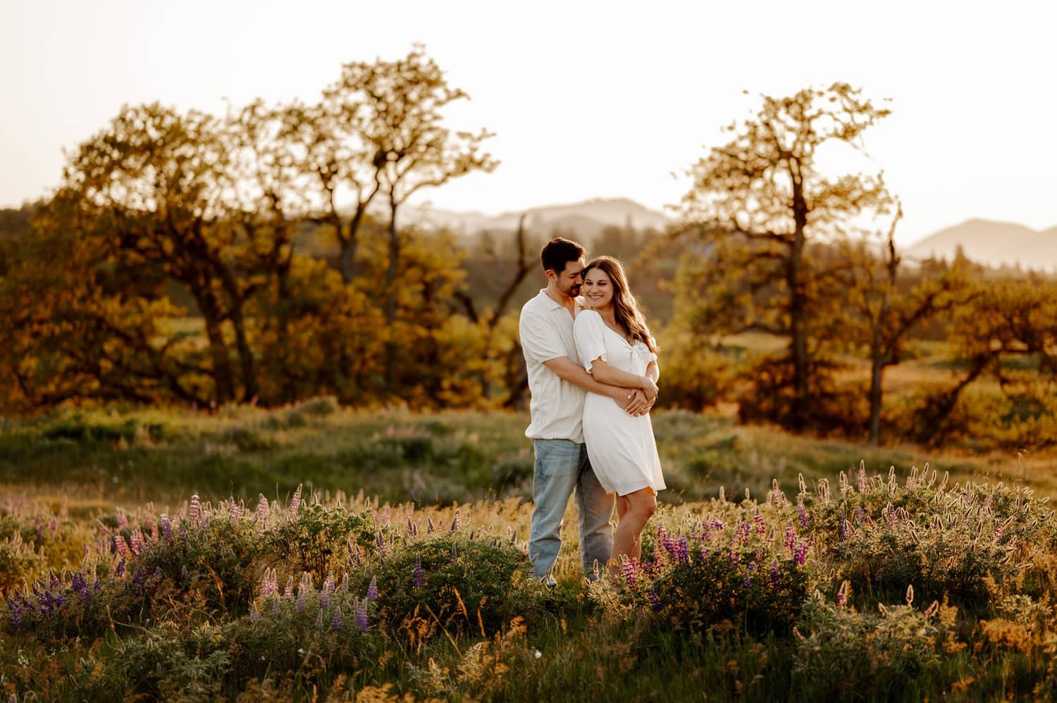 A couple embraces in a field of wildflowers at sunset, surrounded by trees and rolling hills near Rowena Crest during their engagement session.