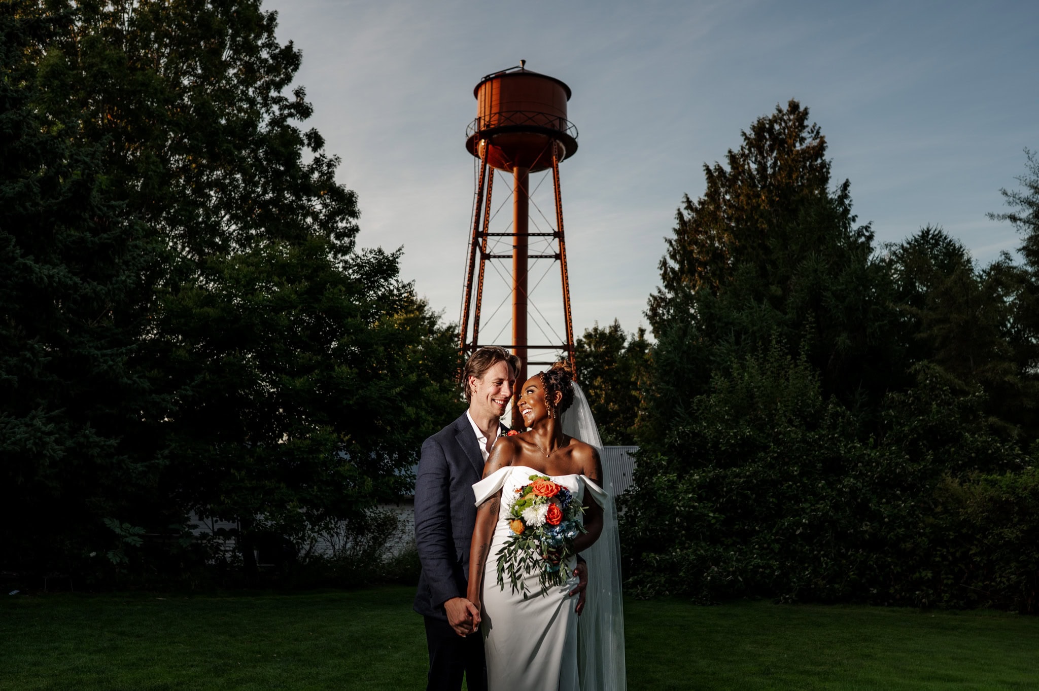 A groom in a suit embraces from behind a bride in a white gown and long veil, standing in front of a waterpower and green trees set against a blue sky at McMenamin's Edgefield's Blackberry Hall, photographed by Kyle Carnes Photography.