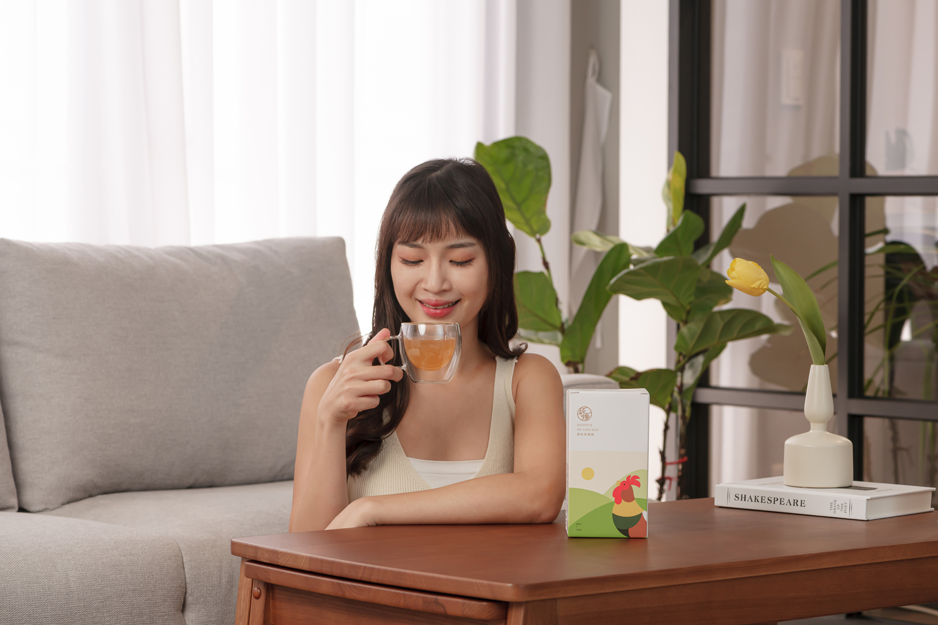 捕捉溫暖午後時光，年輕女性優雅地享受純煉滴雞精 Essence of chicken，木質桌面上置放產品盒與書籍裝飾，窗邊綠植襯托舒適居家氛圍，台中廣告攝影專家奇力克影像工作室 KYRIC STUDIO 精心打造的商業視覺。