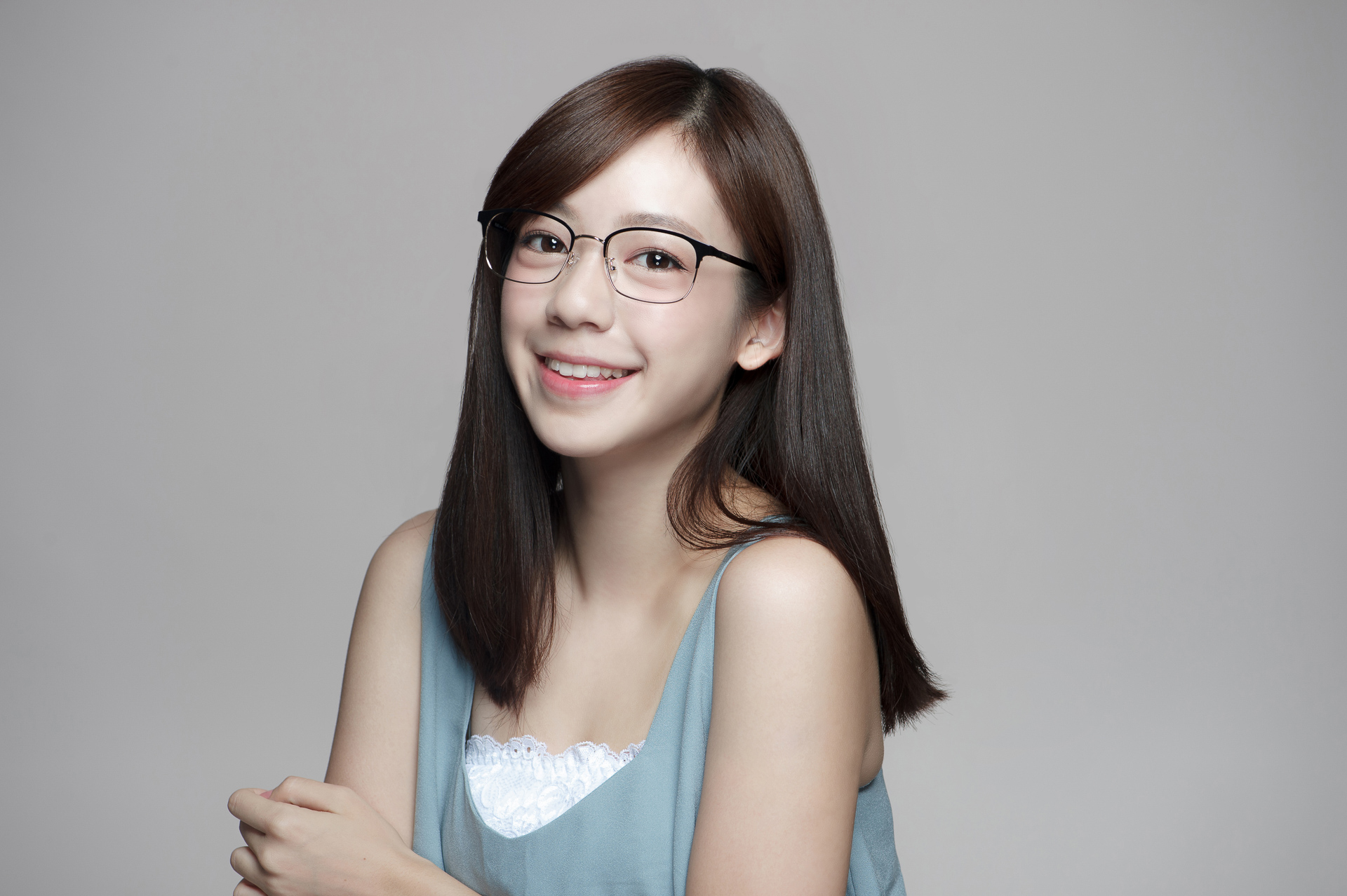 戴著Plume Eyewear眼鏡的年輕女性,露出燦爛笑容,長黑髮披肩,身穿淺藍色上衣,簡潔灰白背景凸顯清爽氣質。中部商業攝影專家奇力克影像工作室KYRIC STUDIO精心打造的專業形象作品。