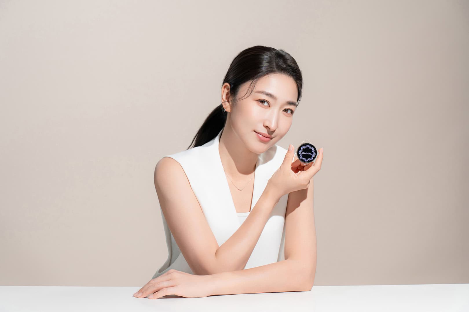 奇力克影像工作室 KYRIC STUDIO 為 Rachel Wine 拍攝的中部美妝攝影，展現亞洲女性持握深藍色唇膏產品，白色無袖上衣搭配簡約頸鍊，柔和打光突顯膚質光澤，中性米灰背景襯托專業質感，構圖優雅呈現品牌形象。