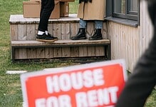 A woman removes a 'House For Rent' sign as new tenants move in, exchanging packages and flowers.