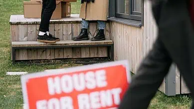A woman removes a 'House For Rent' sign as new tenants move in, exchanging packages and flowers.