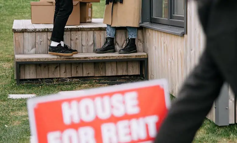 A woman removes a 'House For Rent' sign as new tenants move in, exchanging packages and flowers.