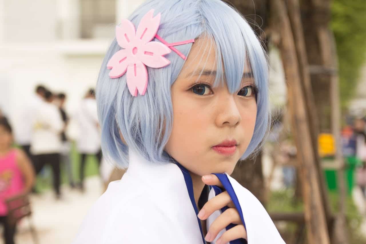 Close-up portrait of a young woman in cosplay with blue hair and floral accessory outdoors.