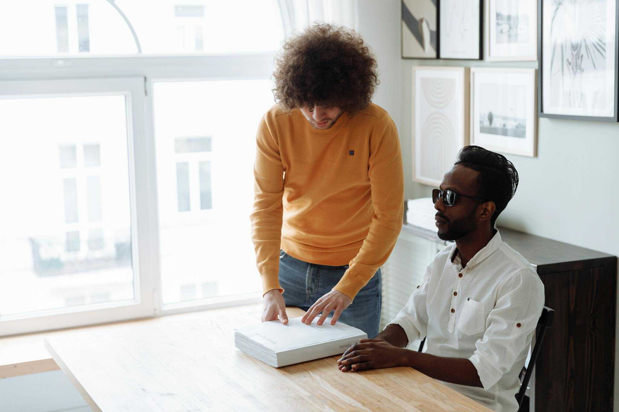 A mentor assists a visually impaired man in reading a Braille book at home.