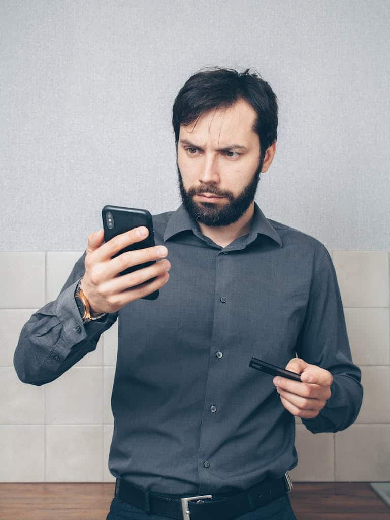 An adult man in a gray shirt looks concerned while using a smartphone and holding a credit card indoors.