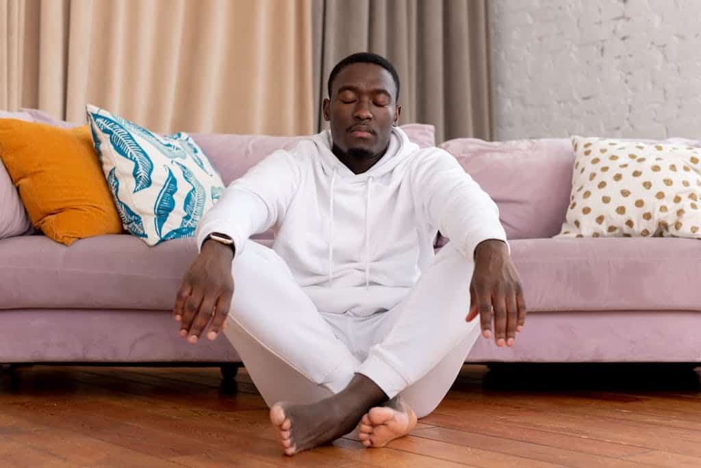 Calm African American male in white clothes sitting with closed eyes and crossed legs on floor against comfortable sofa in living room