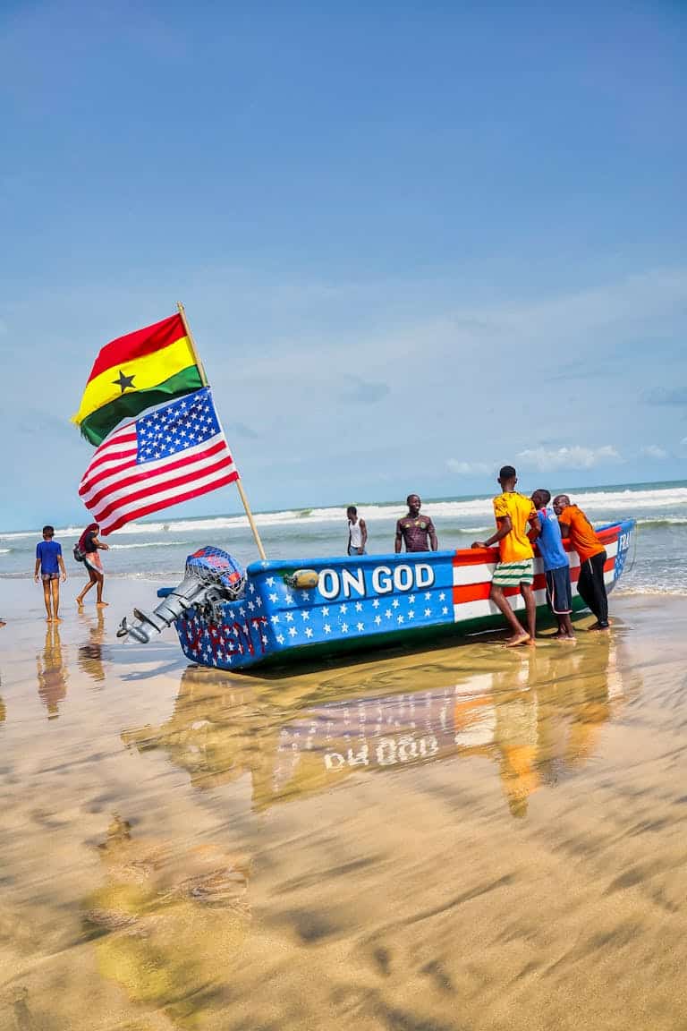 Group of people near a boat with Ghana and USA flags on a sunny beach.