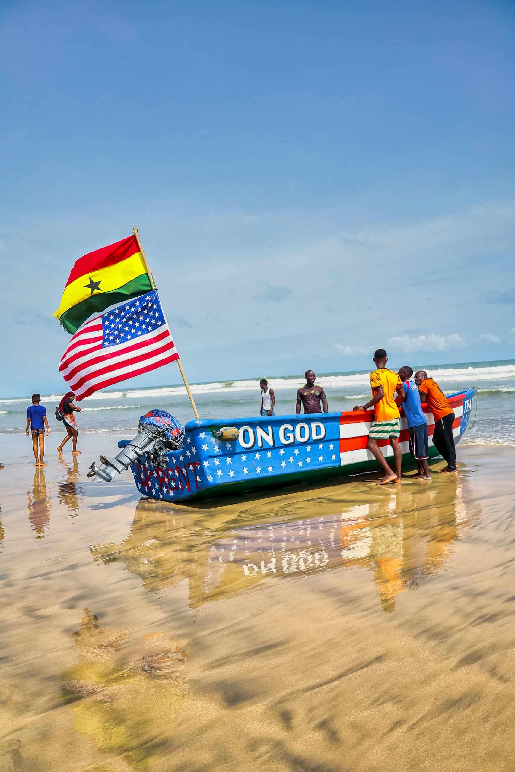 Group of people near a boat with Ghana and USA flags on a sunny beach.