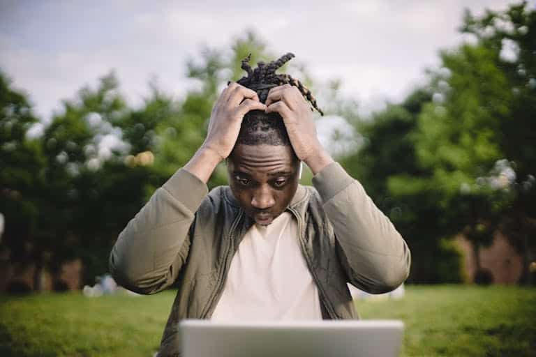 Upset young African American male freelancer clutching head with hands after failure in project while working remotely in park