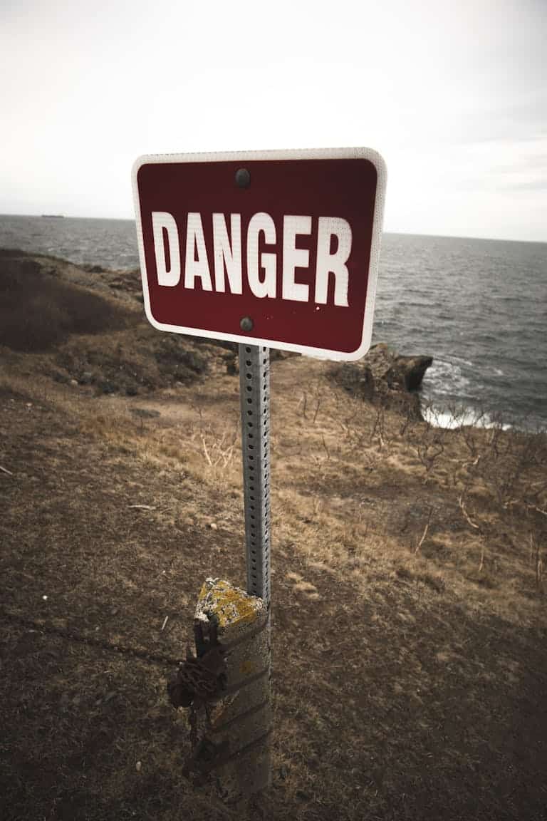 Weathered danger sign by a rugged sea cliff, warning of hazardous terrain.