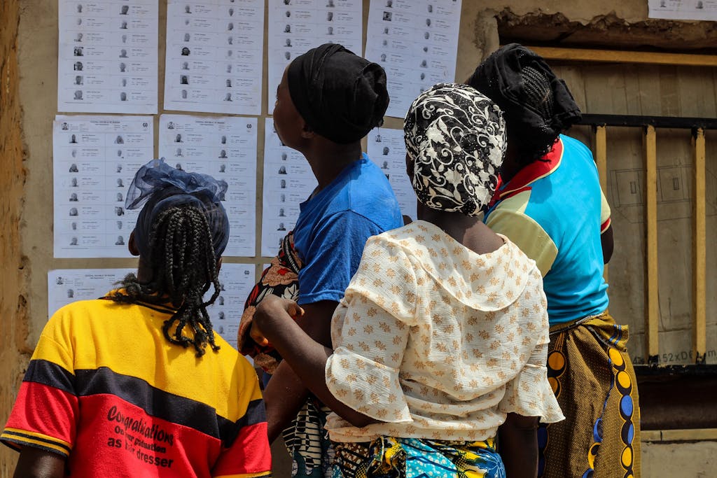 A group of women reading election lists posted on a wall outdoors during the day.