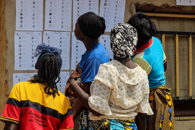 A group of women reading election lists posted on a wall outdoors during the day.