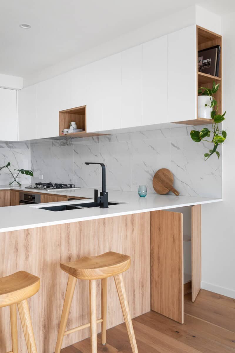 20 Kitchen Splashback Ideas Auckland Homeowners Are Choosing (2026) 2 White marble-look slab splashback running between a white benchtop and overhead cabinets, with a black mixer tap and timber-look base cabinets