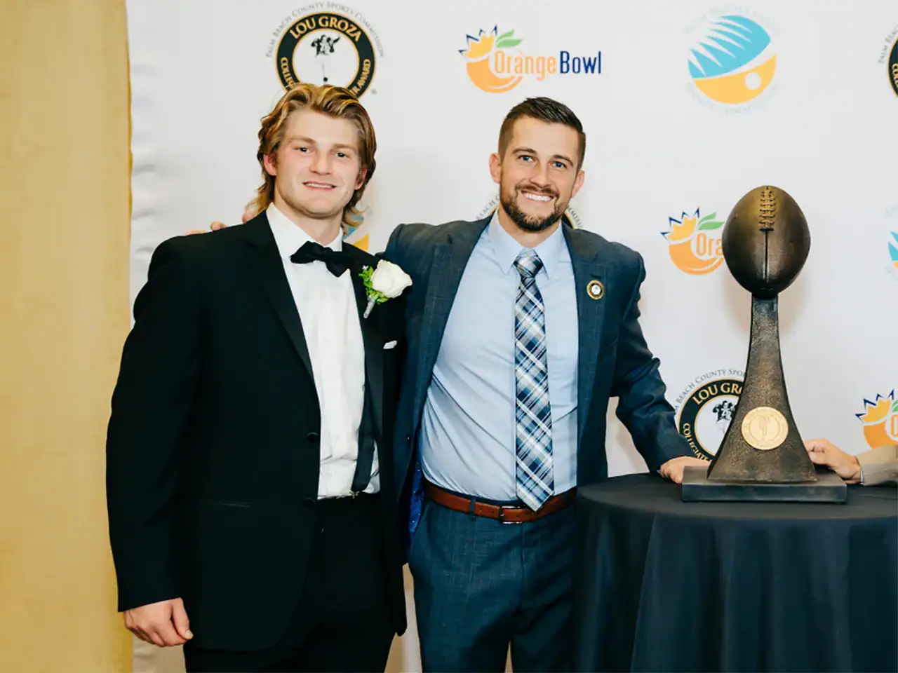 Two men in formal suits at Lou Groza Award event with trophy and backdrop.