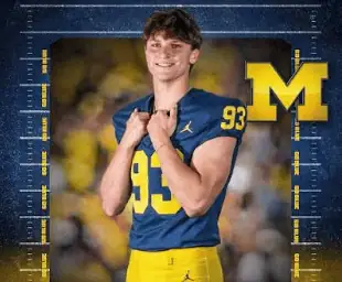Young football player in Michigan Wolverines uniform smiling with M logo in background.